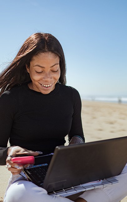A woman sitting on the beach, holding a computer and tablet.
